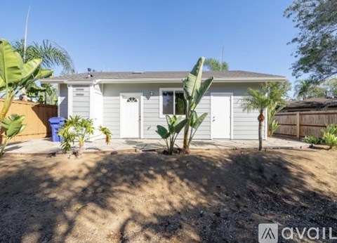 A house with a white exterior and a brown roof is surrounded by a dirt area with plants and trees.