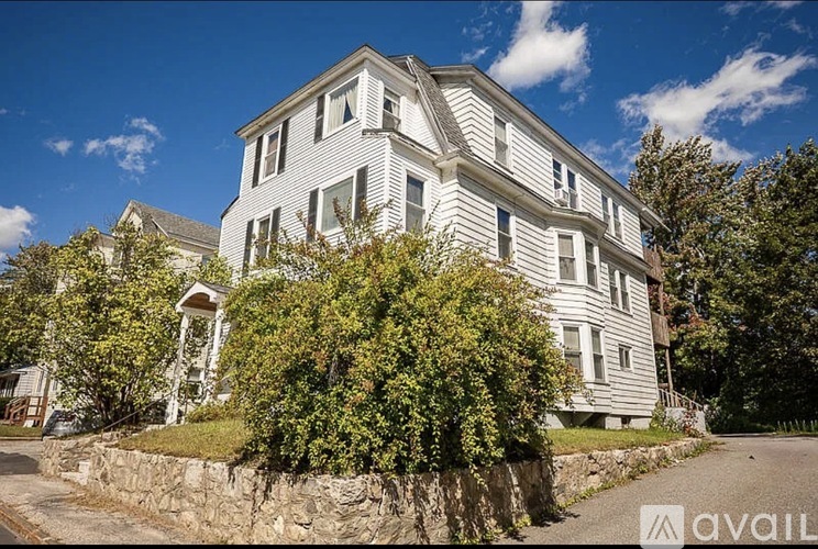 A large, two-story house with a grey exterior and white trim is surrounded by greenery and a stone wall.