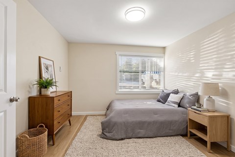 Bedroom with a queen bed, a wood-tone dresser, a nightstand, and a window with blinds on plank-style flooring.
