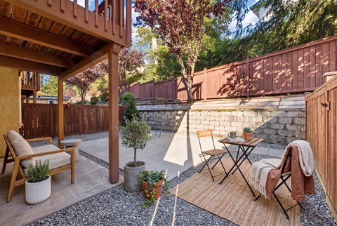 Private ground-level patio with a cedar fence, stone retaining wall, lounge chair, and bistro table under a wood deck.