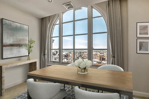 A dining room with a wooden table and chairs, a vase with white flowers, and a large window with a view of the city.
