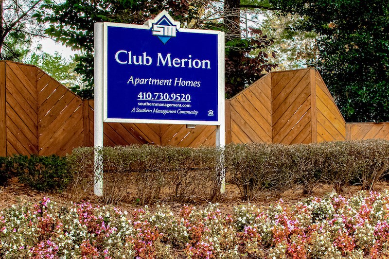 a sign for club merion apartments in front of a fence and flowers