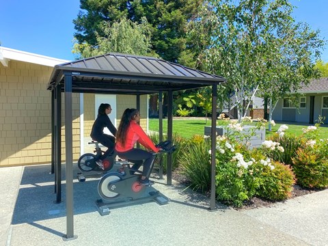 Two people are exercising on a stationary bike under a shelter.