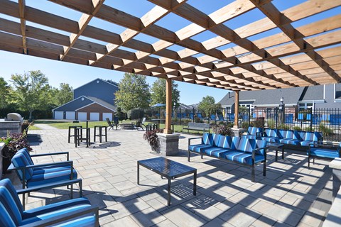 A wooden pergola shades a seating area with blue chairs and a table.