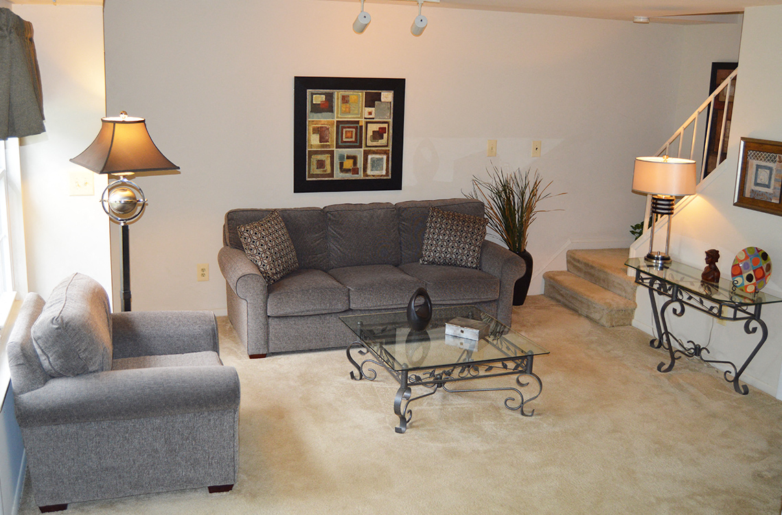 A living room with a grey couch, a grey chair, a glass coffee table and a painting on the wall.