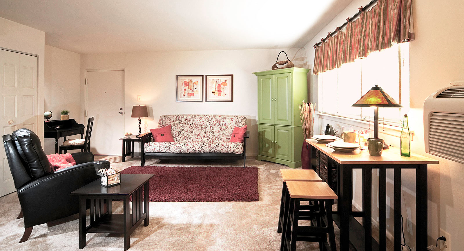 A living room with a black leather chair, a patterned sofa, a small table with a lamp, and a green cabinet.