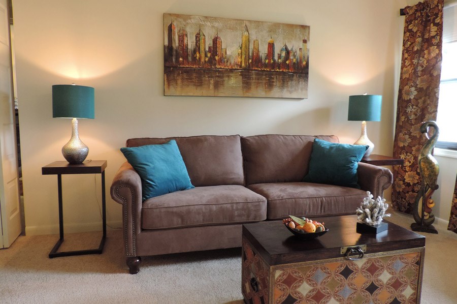 A brown couch with blue pillows sits in a living room with a painting on the wall.