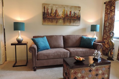 A brown couch with blue pillows sits in a living room with a painting on the wall.