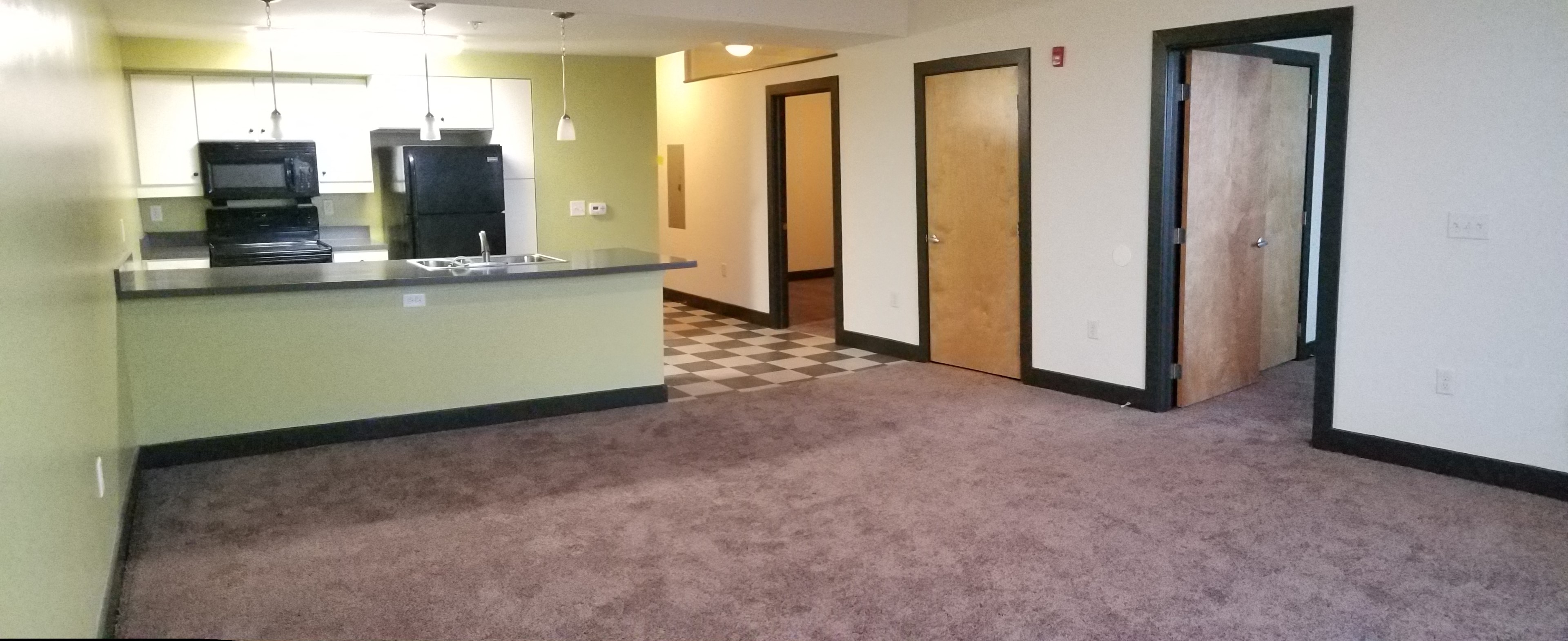 an empty living room with a carpeted floor and a kitchen with doors