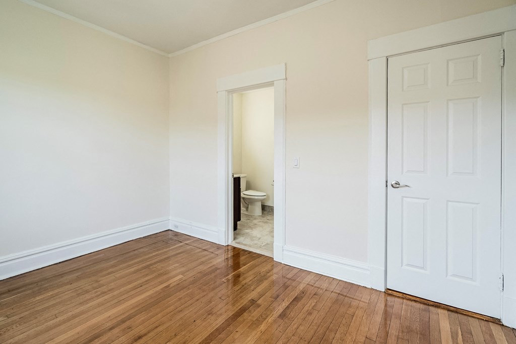 a bedroom with white walls and wood flooring and a door to a bathroom