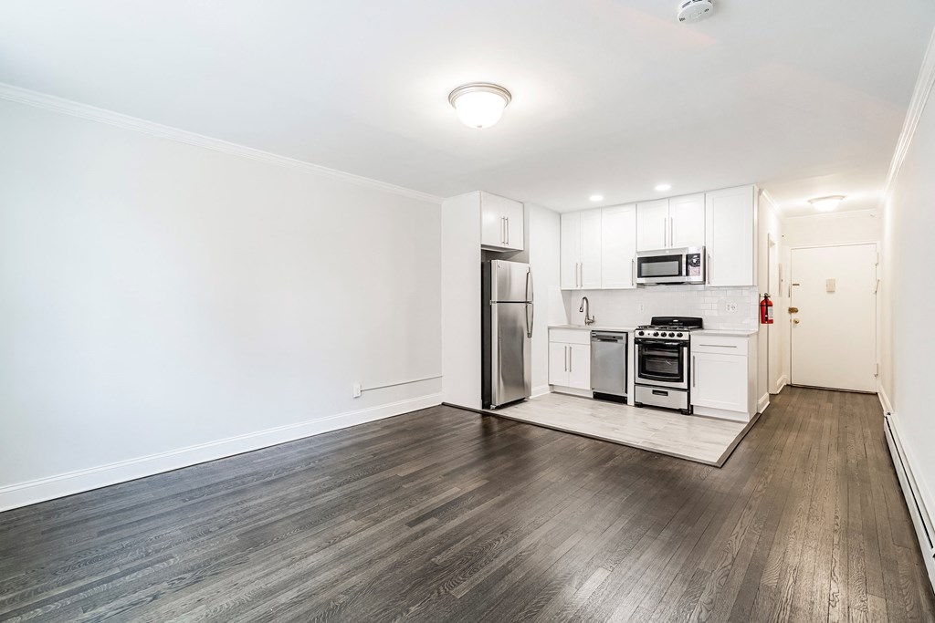 a living room with white walls and wooden floors and a kitchen with stainless steel appliances