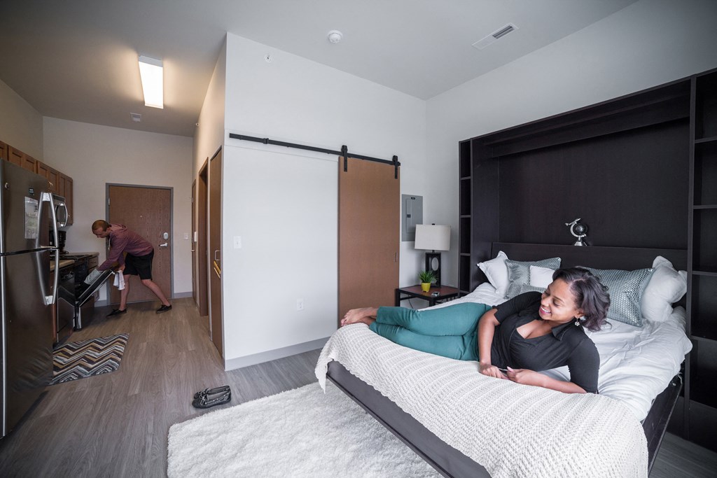 a woman laying on a bed in a bedroom with a man working in a kitchen