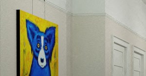 A woman is sitting on a couch in a living room with a ceiling fan and a painting of a dog on the wall.