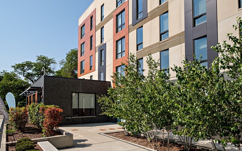 A modern building with a red and beige facade is surrounded by greenery.