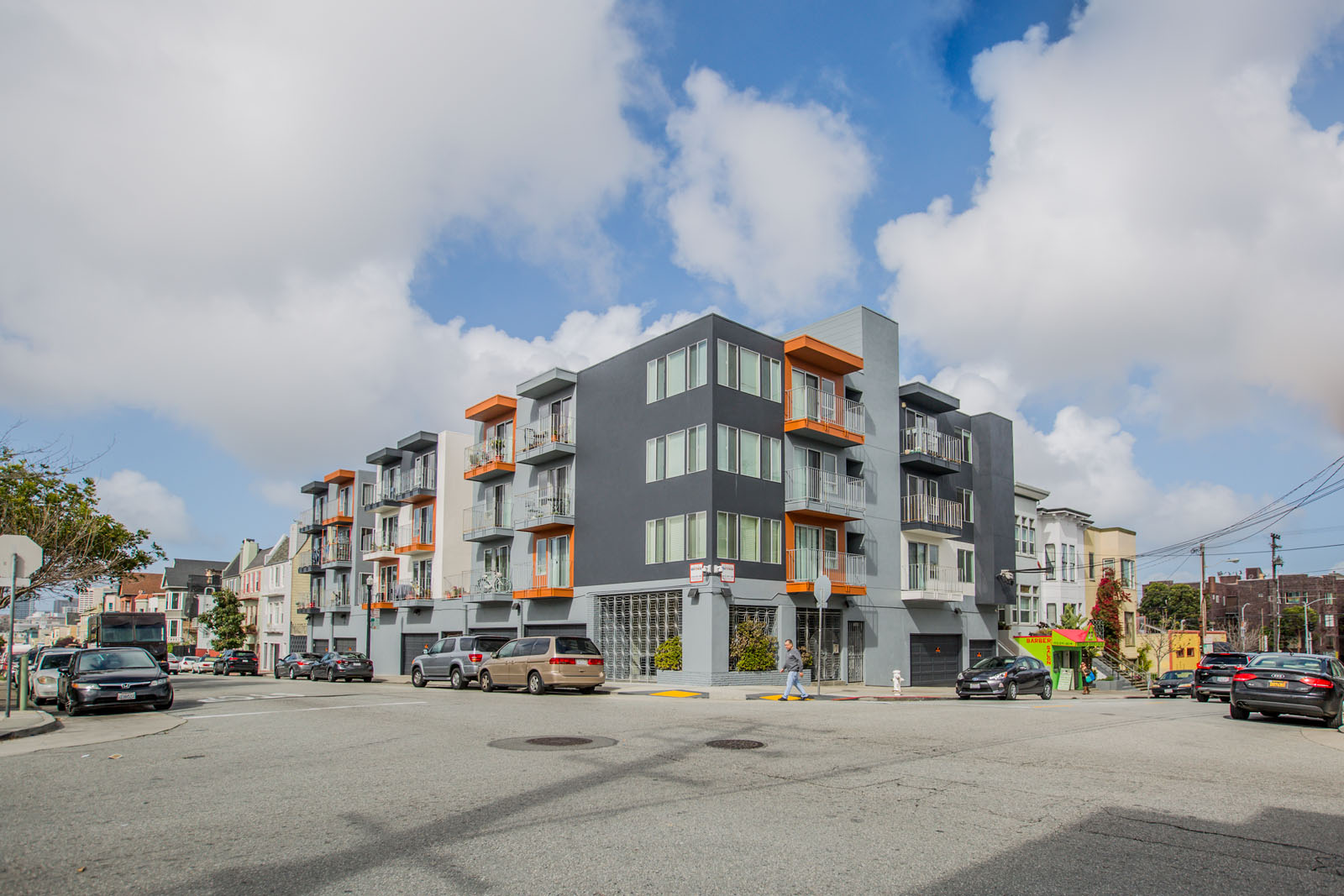 A modern multi-story building with orange accents is situated on a street corner with cars parked in front.