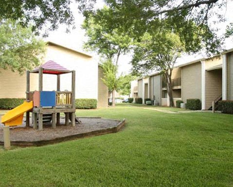 A playground with a slide and a wooden structure is surrounded by a grassy area and apartment buildings.