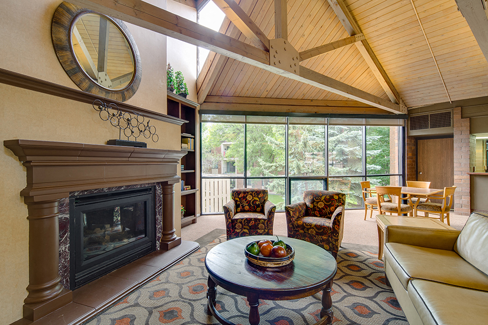 Community room with chairs facing fireplace, high ceilings with wooden archways, and large windows