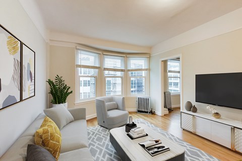 A living room with a grey sofa, a white coffee table, a grey armchair, a white TV stand, a potted plant, and a painting on the wall.