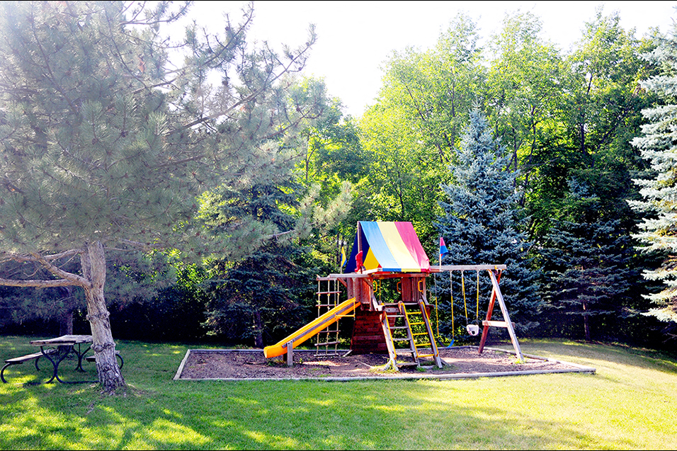 outdoor playground with slide and swing next to a park bench and large trees all around