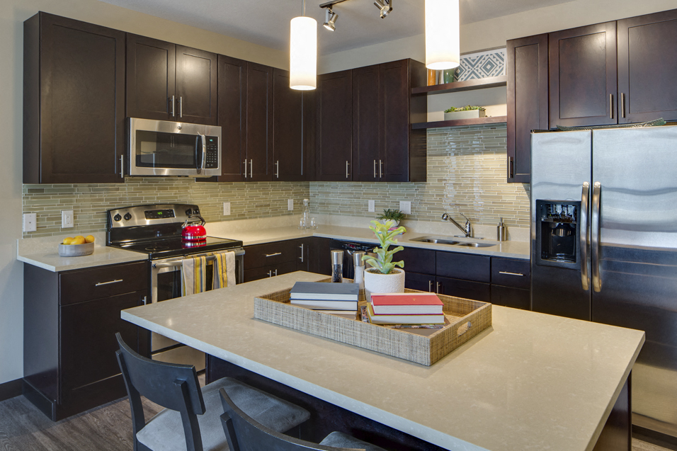 Kitchen with modern appliances, white countertops, brown cabinets, and an island with books and a plant on it