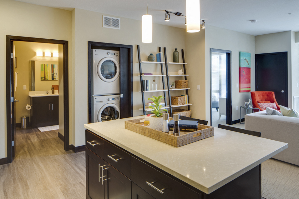 Wide shot of apartment. Visible bathroom, washer and dryer, kitchen island, and living room couch
