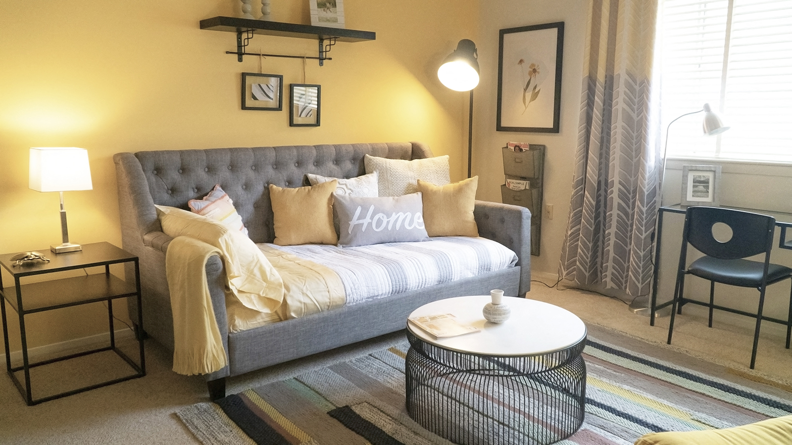 A grey tufted sofa with a white throw blanket sits in a living room with a black coffee table in front of it.