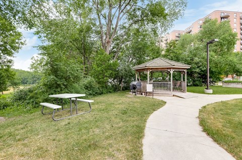 A gazebo sits in the middle of a grassy area with a picnic table and a concrete walkway.