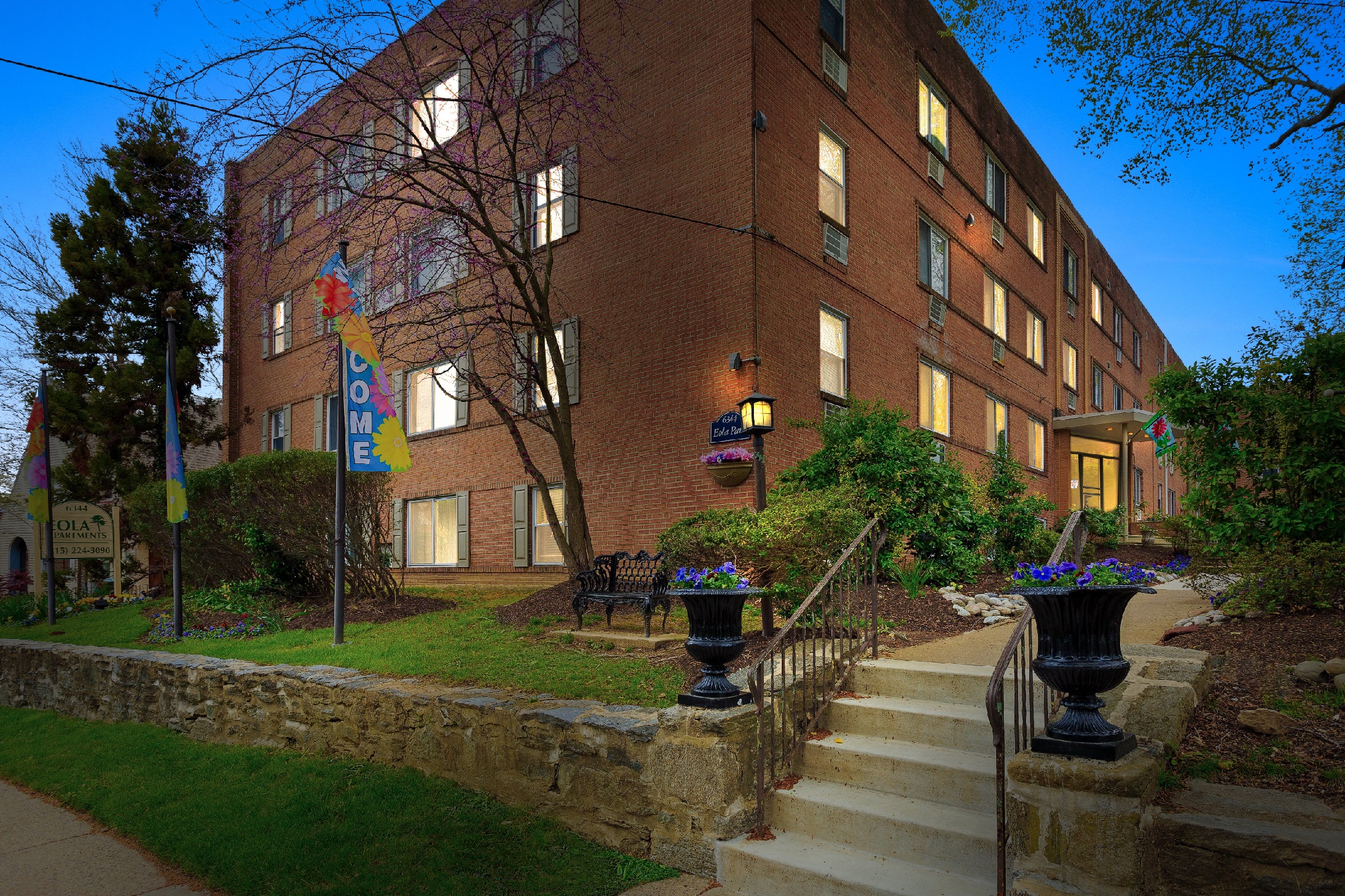 Exterior of building at night, showing landscaped grass, flags, garden, and stairs and walkway to building entrance.
