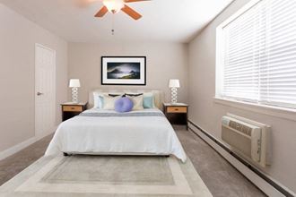Furnished model master bedroom with ceiling fan and two windows over an air conditioner.