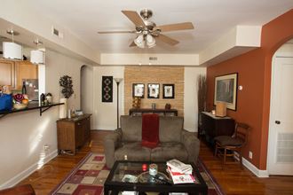 Furnished model living room with ceiling fan, red and orange walls, and wood tile flooring. Has window that opens to kitchen.
