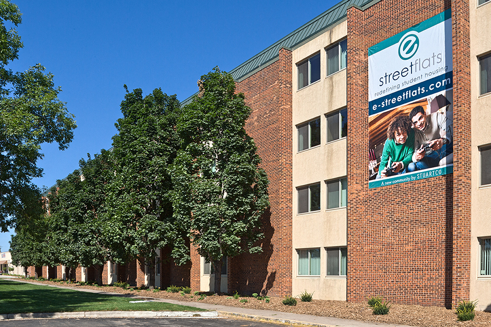outdoor view of property with tan and brown bricks, tall green trees, and a sign on the building that says "e street flats"