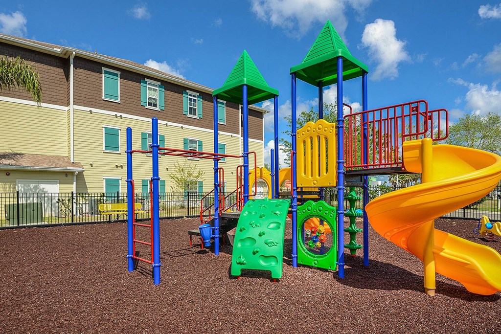 A playground with a yellow slide, green and red play structures, and a brown ground at Cabana Club Apartments, Jacksonville, FL, 32256
