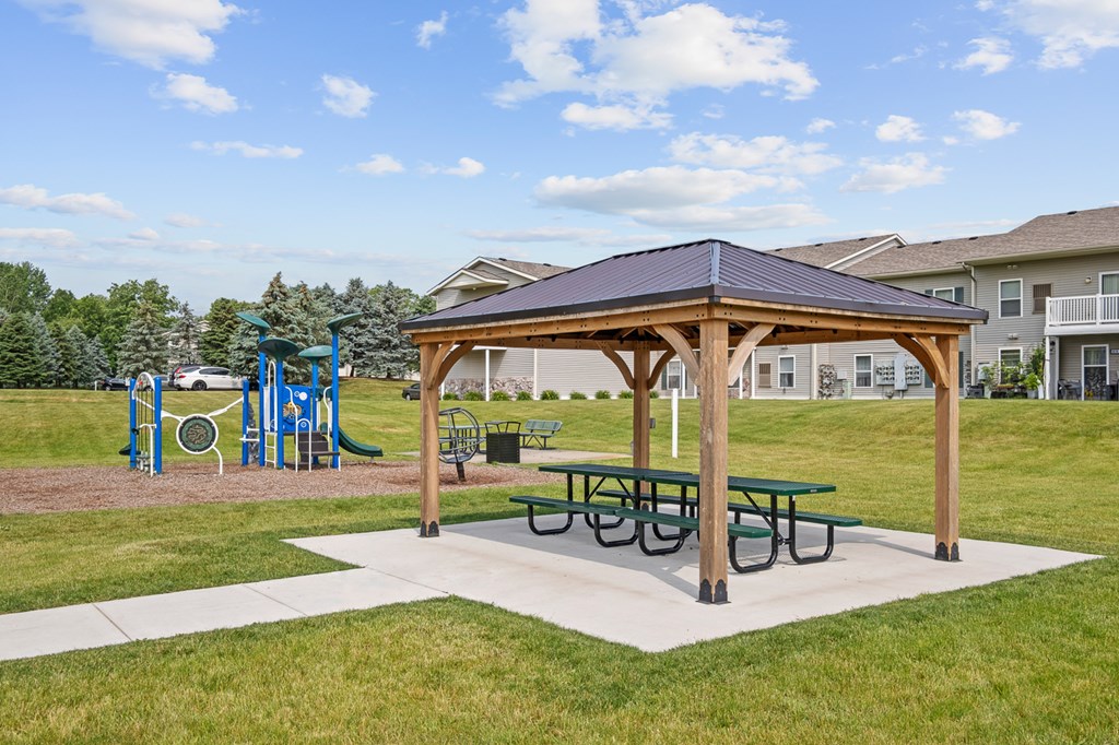 A wooden pavilion with a blue roof and picnic tables sits in a grassy area.