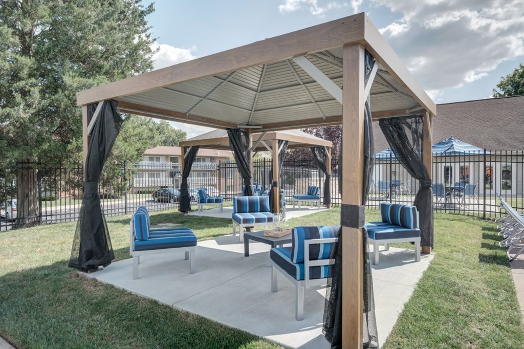 A wooden pavilion with blue cushioned seating is surrounded by a grassy area.