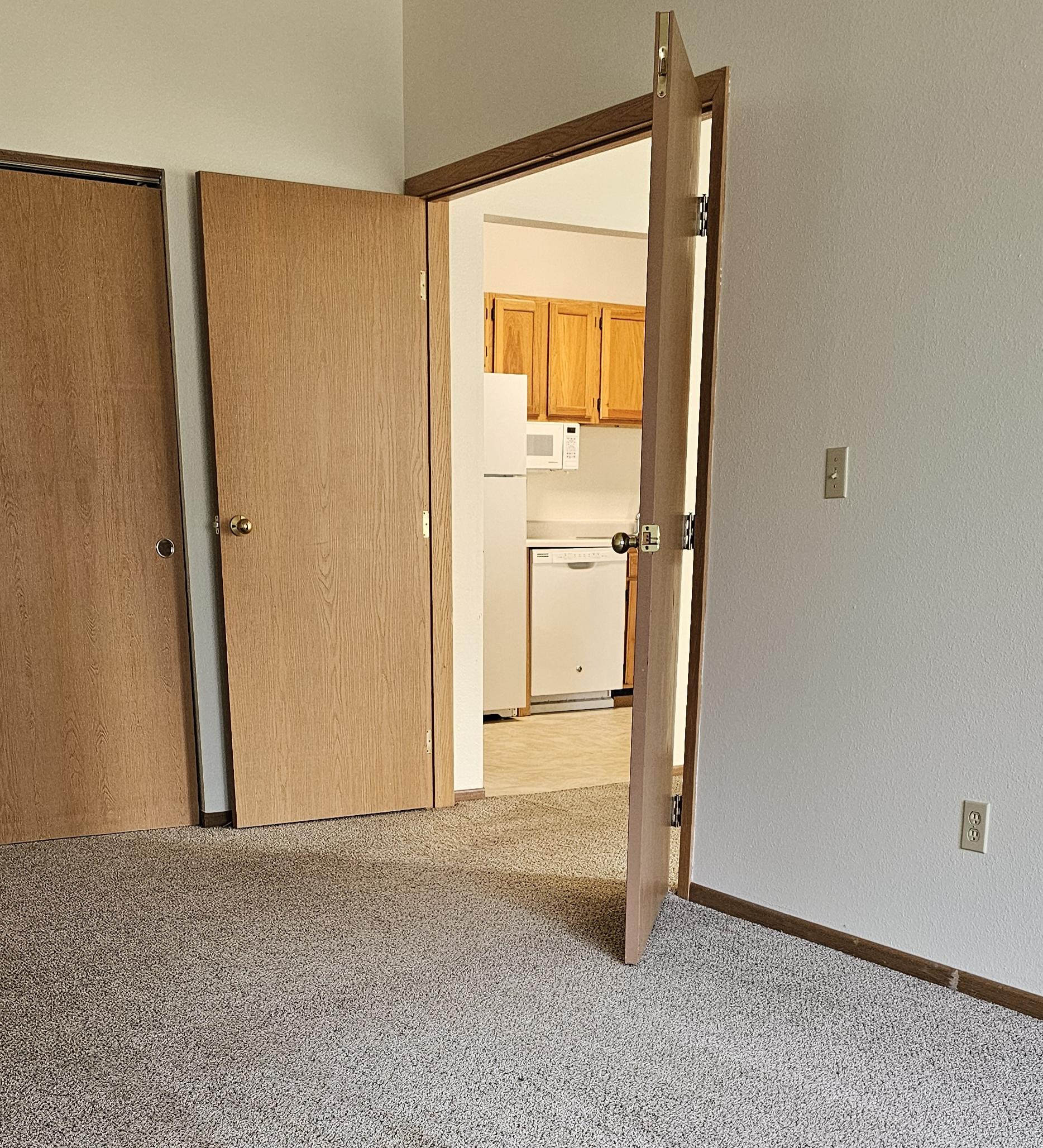 A room with a carpeted floor and a white wall with a doorway leading to a kitchen.