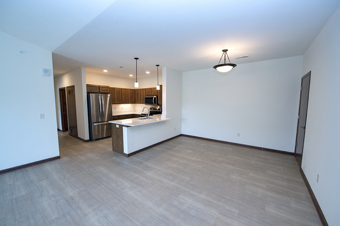 an empty living room with a kitchen and a stainless steel refrigerator