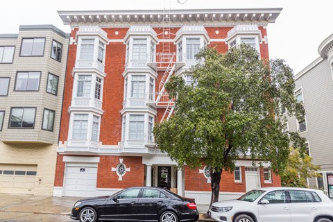 A black car is parked in front of a red brick building with a tree in front of it.