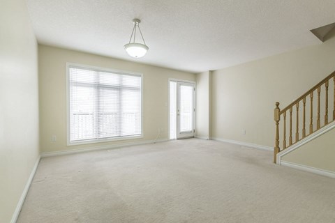 A spacious room with a carpeted floor, a staircase with wooden railings, and two windows with blinds.