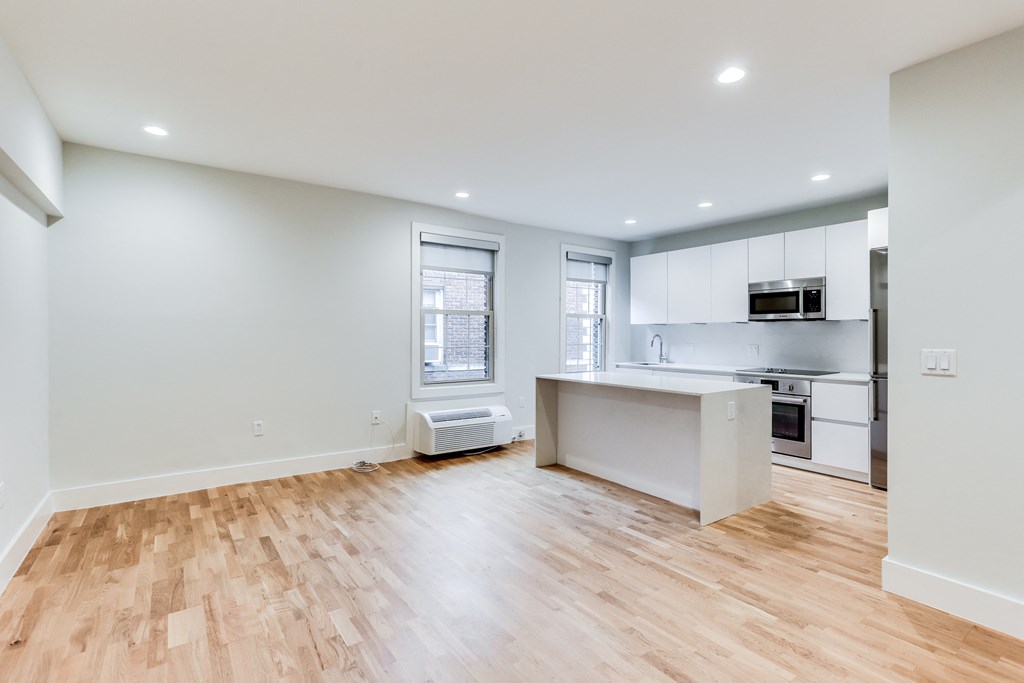 an empty living room with a kitchen with white cabinets and wood floors