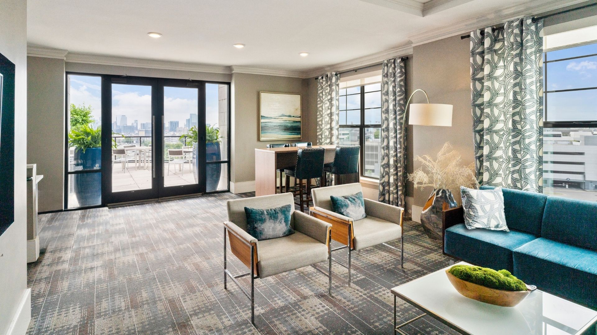 A living room with a grey carpet and a large window overlooking the city.