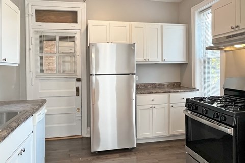 A white refrigerator stands in a kitchen with white cabinets and a black stove.
