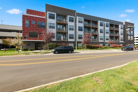 A black car is parked on the side of a road in front of a multi-story apartment building.
