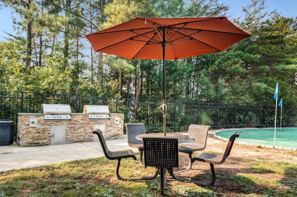 A table with chairs and an umbrella is in the foreground of a pool area.