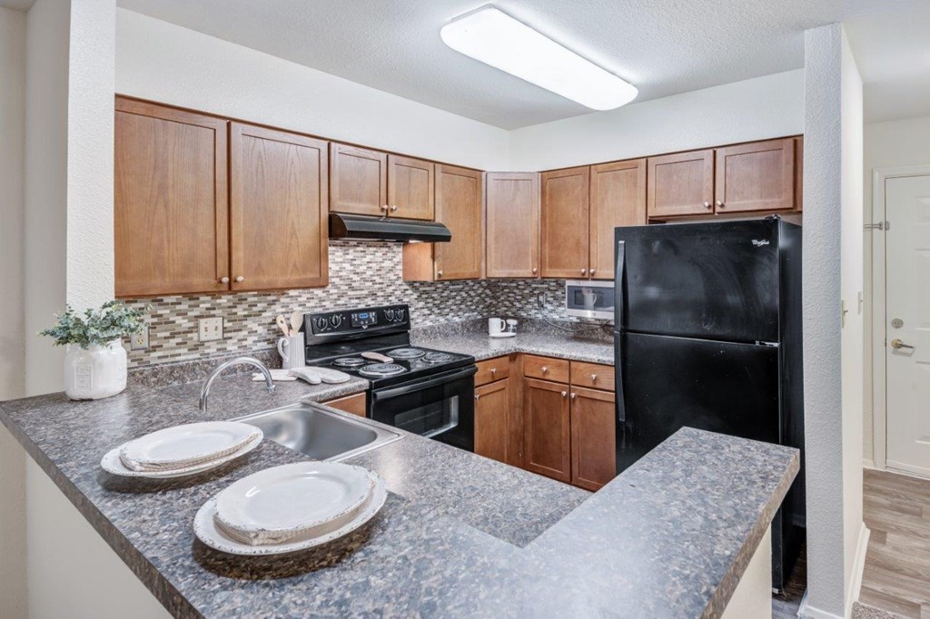 A kitchen with a black refrigerator, brown cabinets, and a marble countertop.