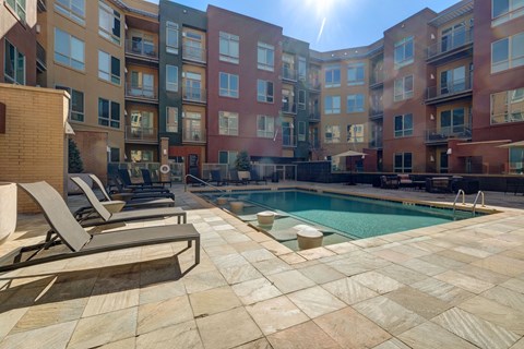 A sunny day at a poolside with lounge chairs and apartment buildings in the background.