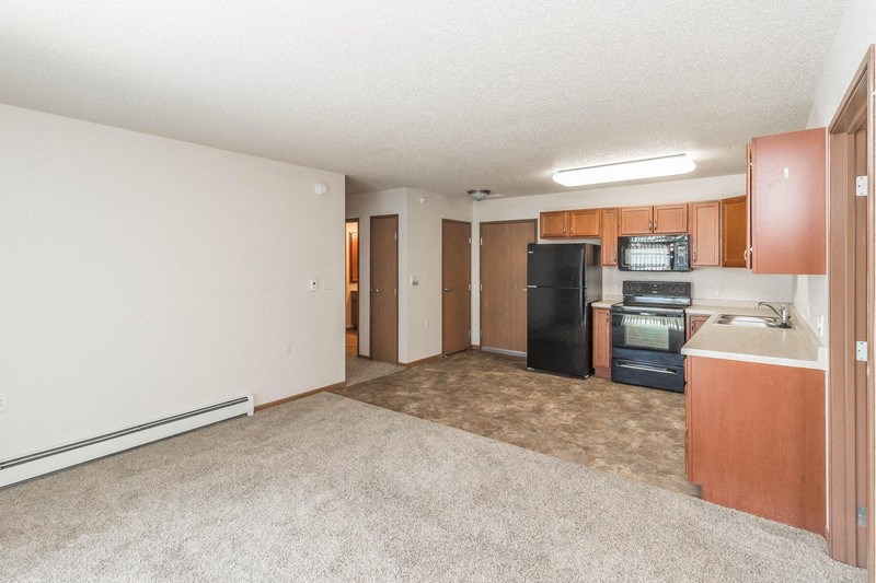 an empty living room with a kitchen with a black refrigerator