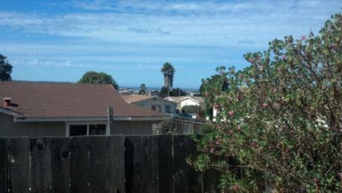 A house with a brown roof is seen from behind a wooden fence with green trees in the background.
