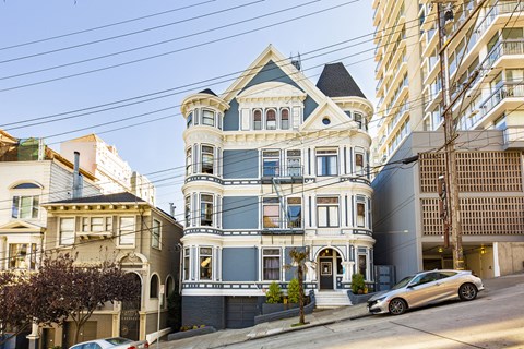 A large, ornate Victorian-style house sits on a street corner with a modern building in the background.