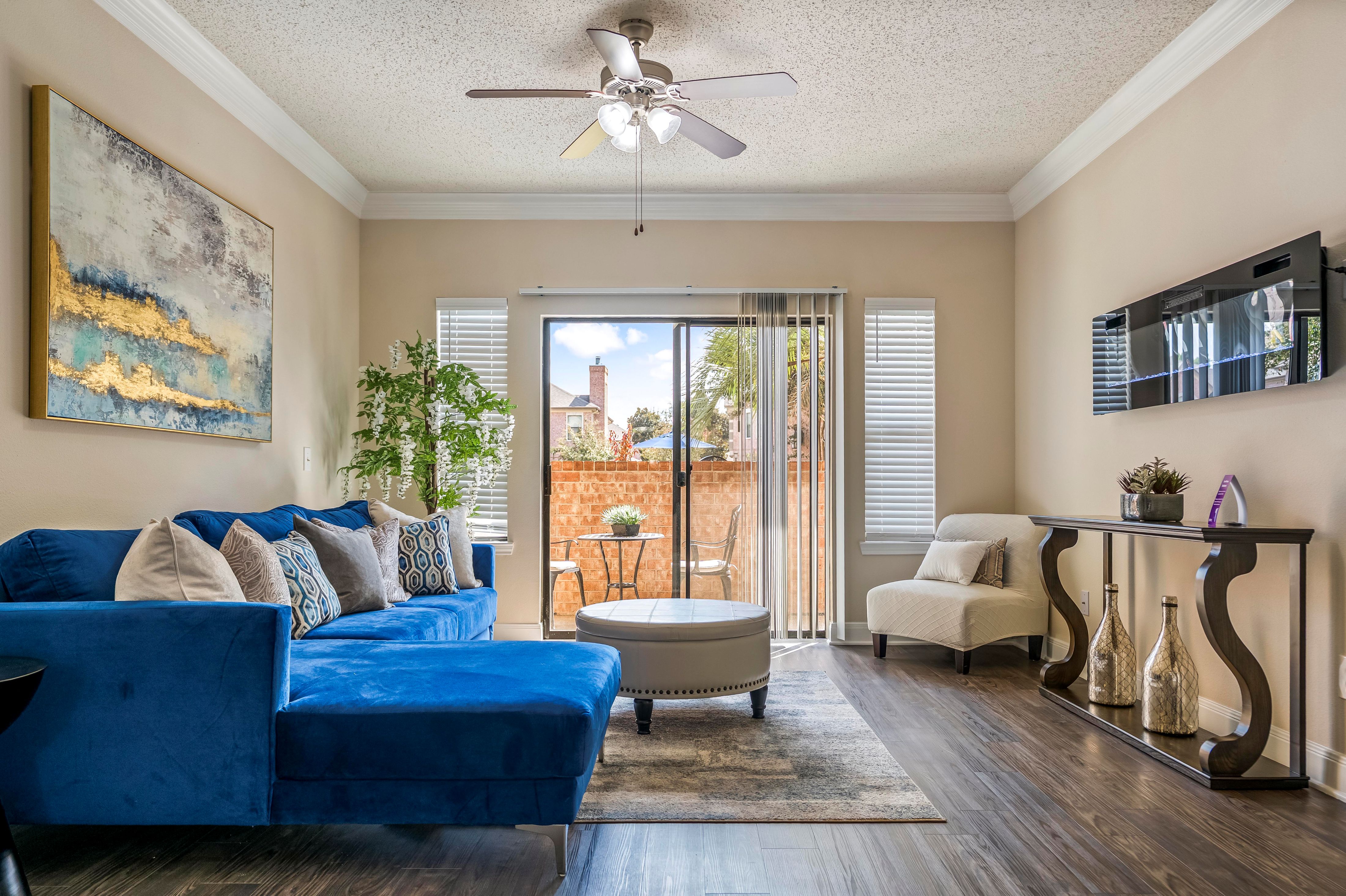 A blue couch sits in a living room with a painting on the wall and a ceiling fan.