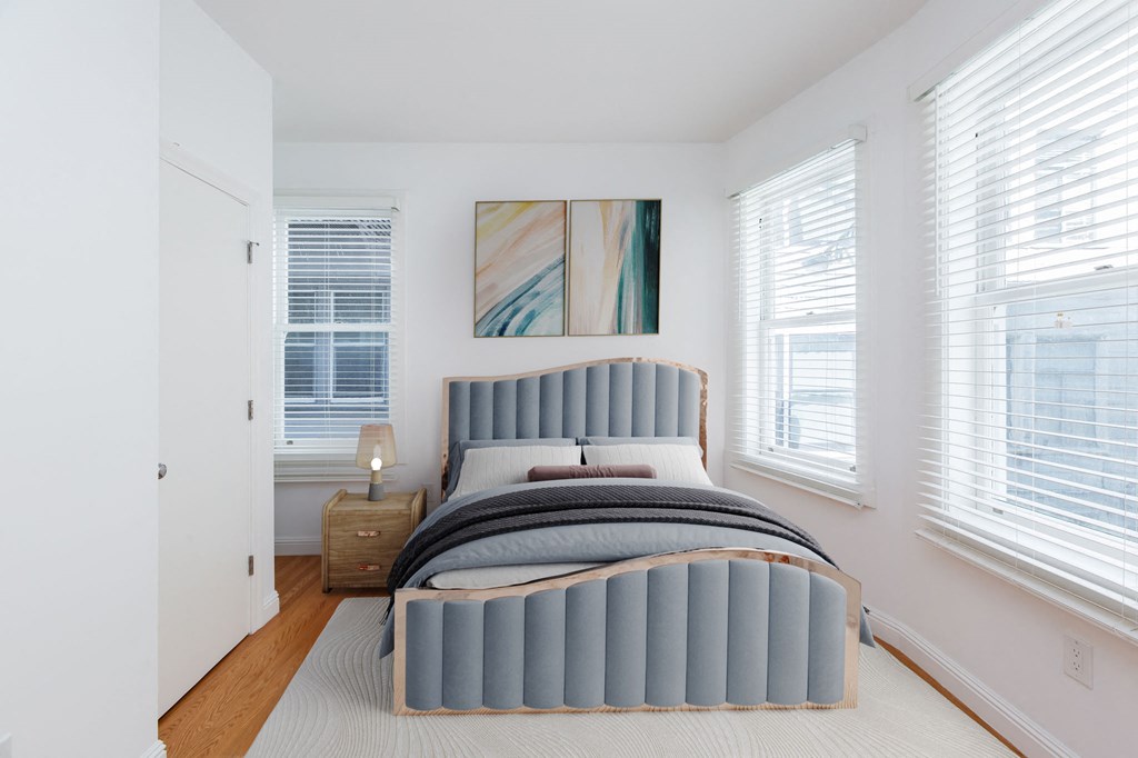 A bed with a grey headboard and pillows is in a room with white walls and a painting hanging above it.
